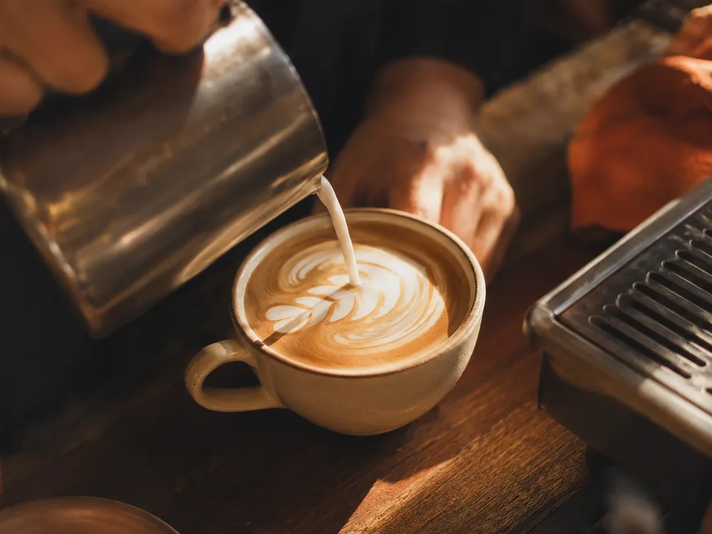 Barista pouring latte art into a flat white, warm morning light on the bench.