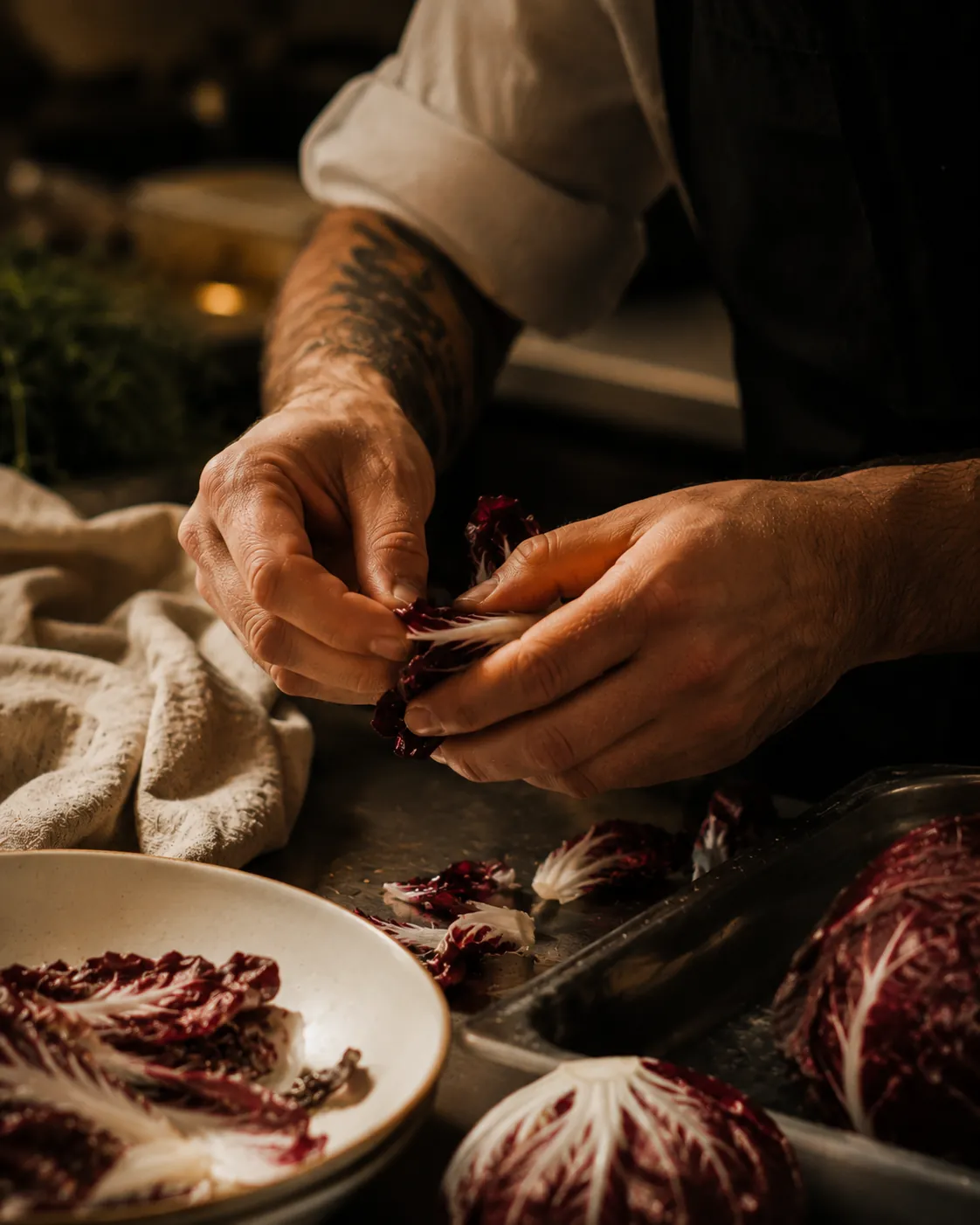 Tattooed chef's hands tearing radicchio over a prep bench in warm kitchen light
