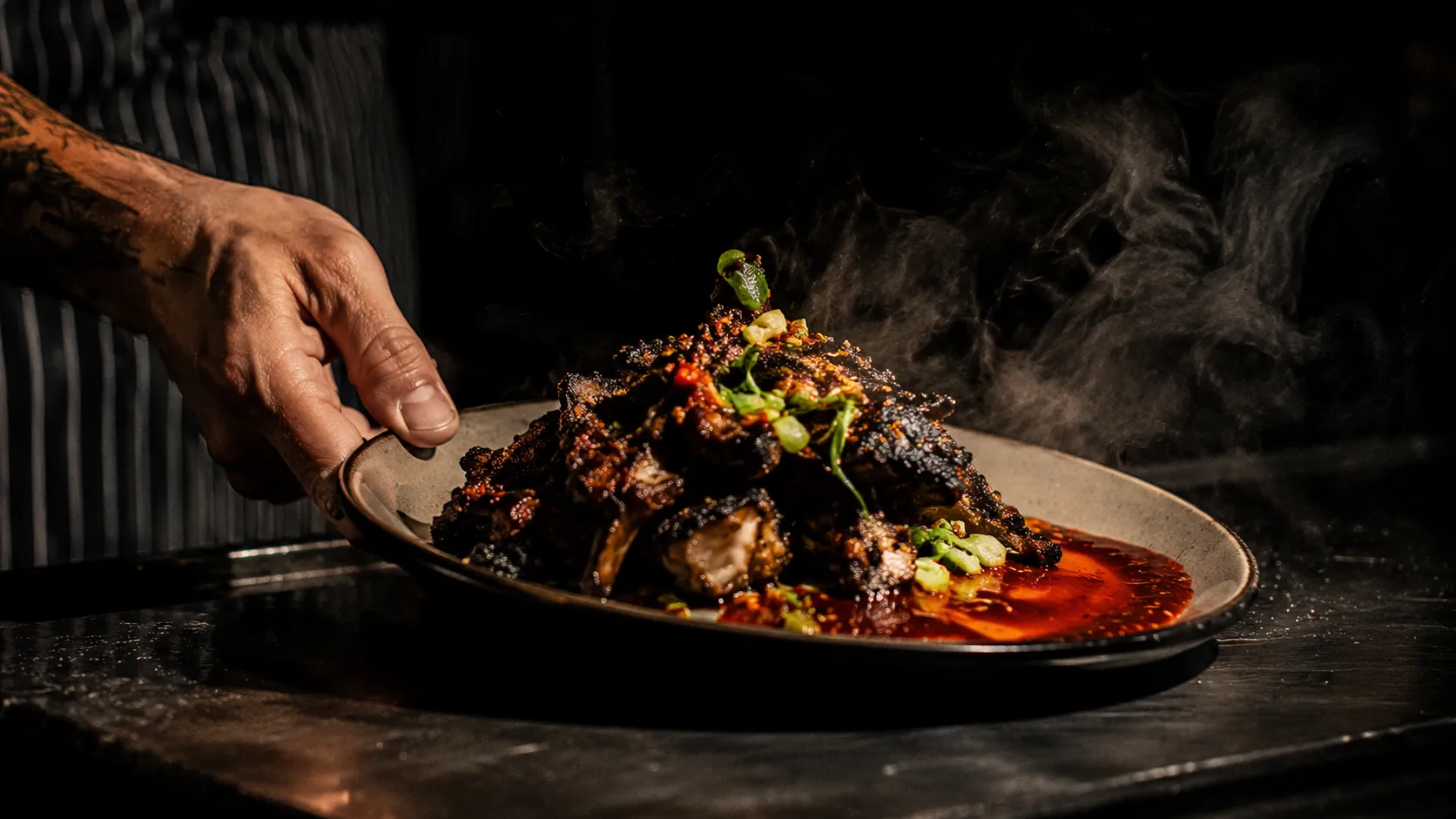 Signature dish arriving on the pass, hard light, steam rising, sauce drizzle mid-plate