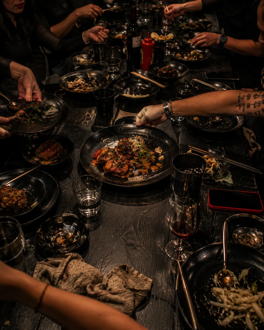 Communal table mid-meal, hands reaching across shared plates, sauce drag marks, wine glasses and napkins in use.